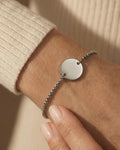 Silver bracelet with a round pendant on a wrist against a beige background